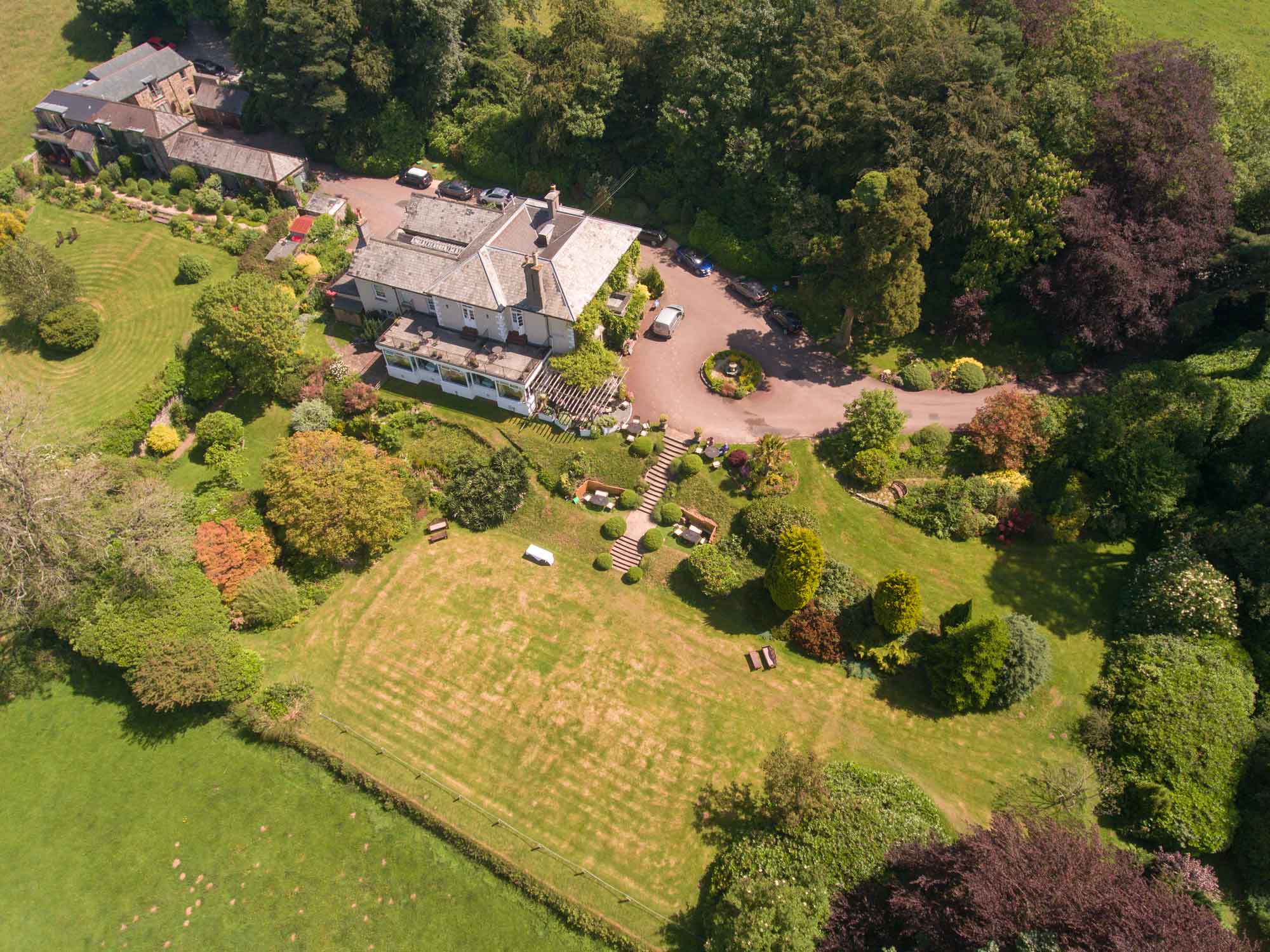 Overhead aerial of a white country hotel with a slate roof and glazed conservatory, its circular gravel drive and parked cars, surrounded by mature trees and terraced lawns