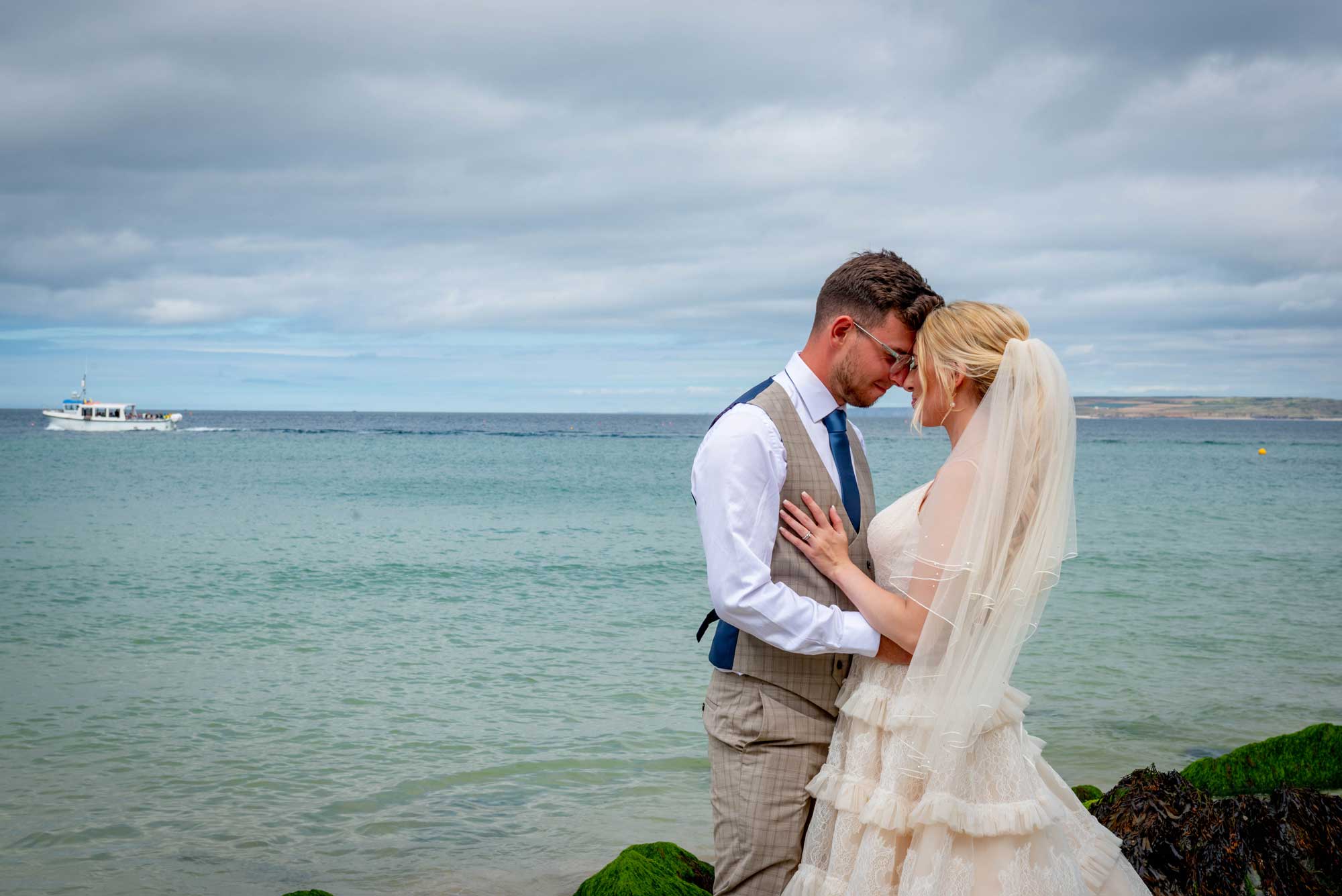 Bride in an ivory lace dress and veil and groom in a checked waistcoat stand forehead to forehead on rocks above a turquoise Cornish bay
